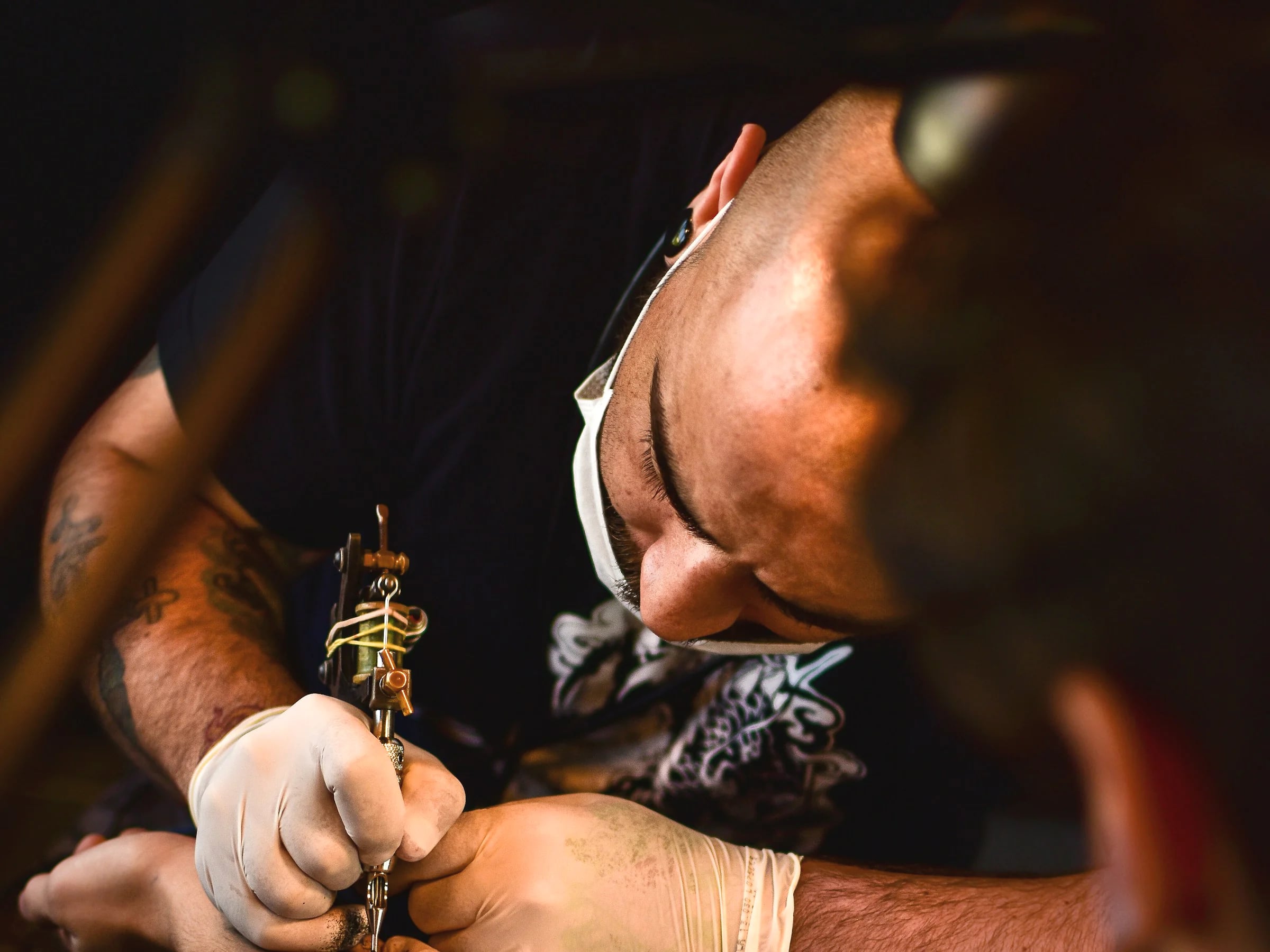 A tattoo artist leaning in to work on a client’s forearm.